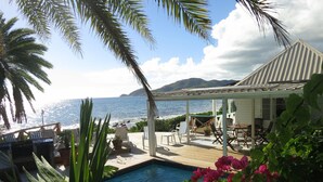 A pool area with a view of the ocean, surrounded by palm trees and a covered patio with outdoor furniture.