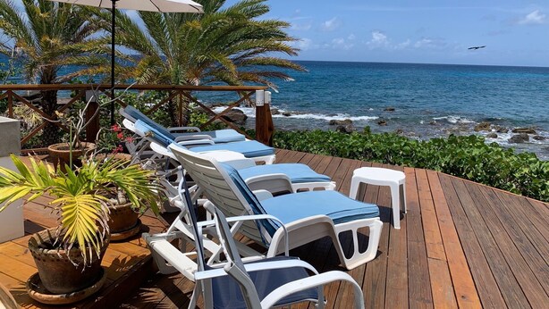 A wooden deck with lounge chairs and a white umbrella, overlooking the ocean with palm trees and a rocky shoreline.