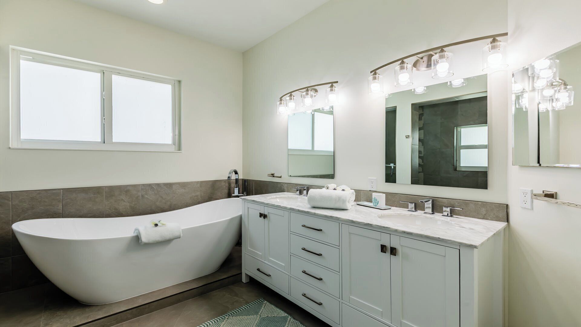 A modern bathroom with a freestanding bathtub, a vanity with a large mirror, and a window above the tub.