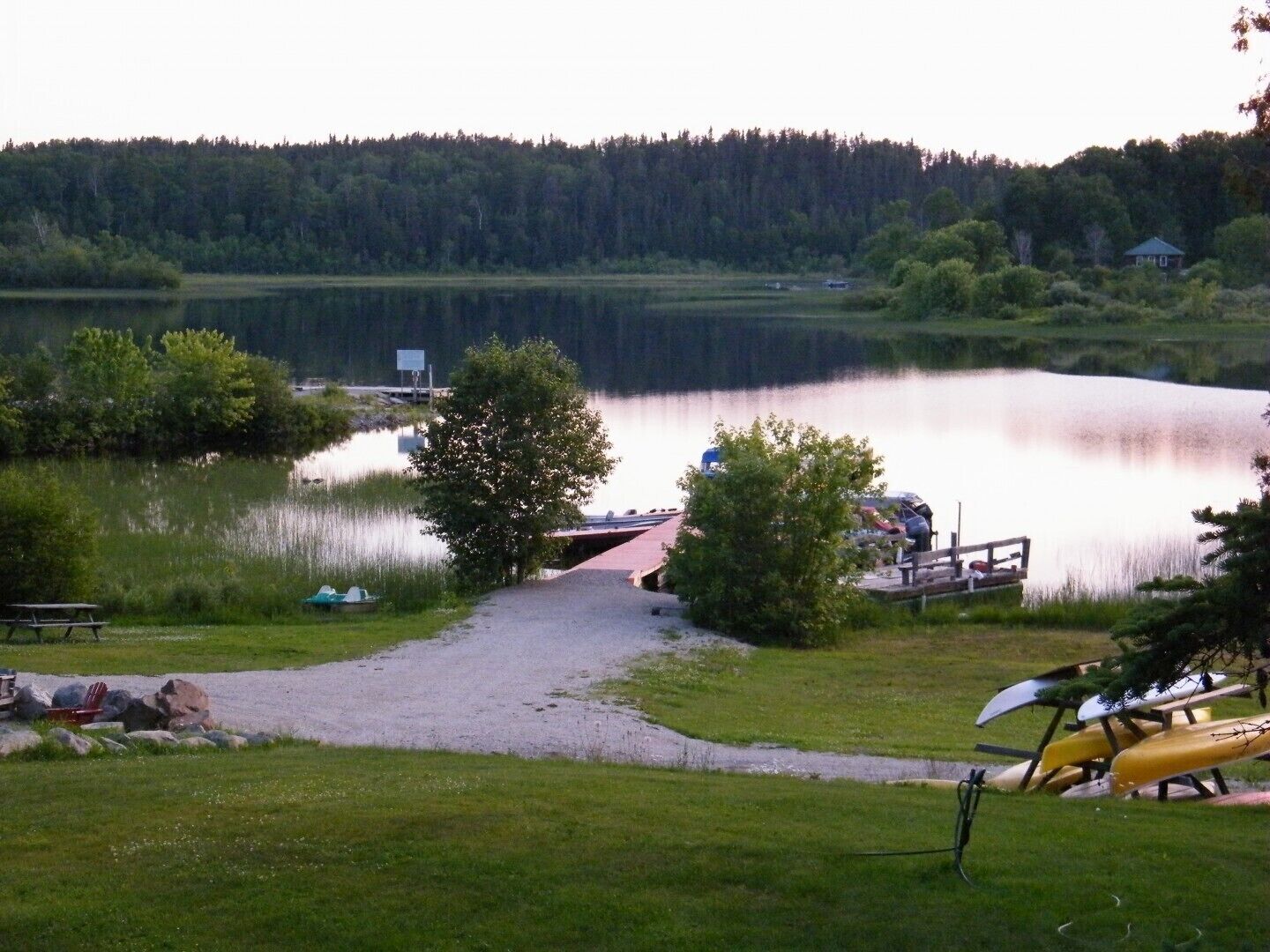 Un terrain de camping au bord du lac, avec un sentier de gravier menant à un quai, des tables de pique-nique et un canoë.