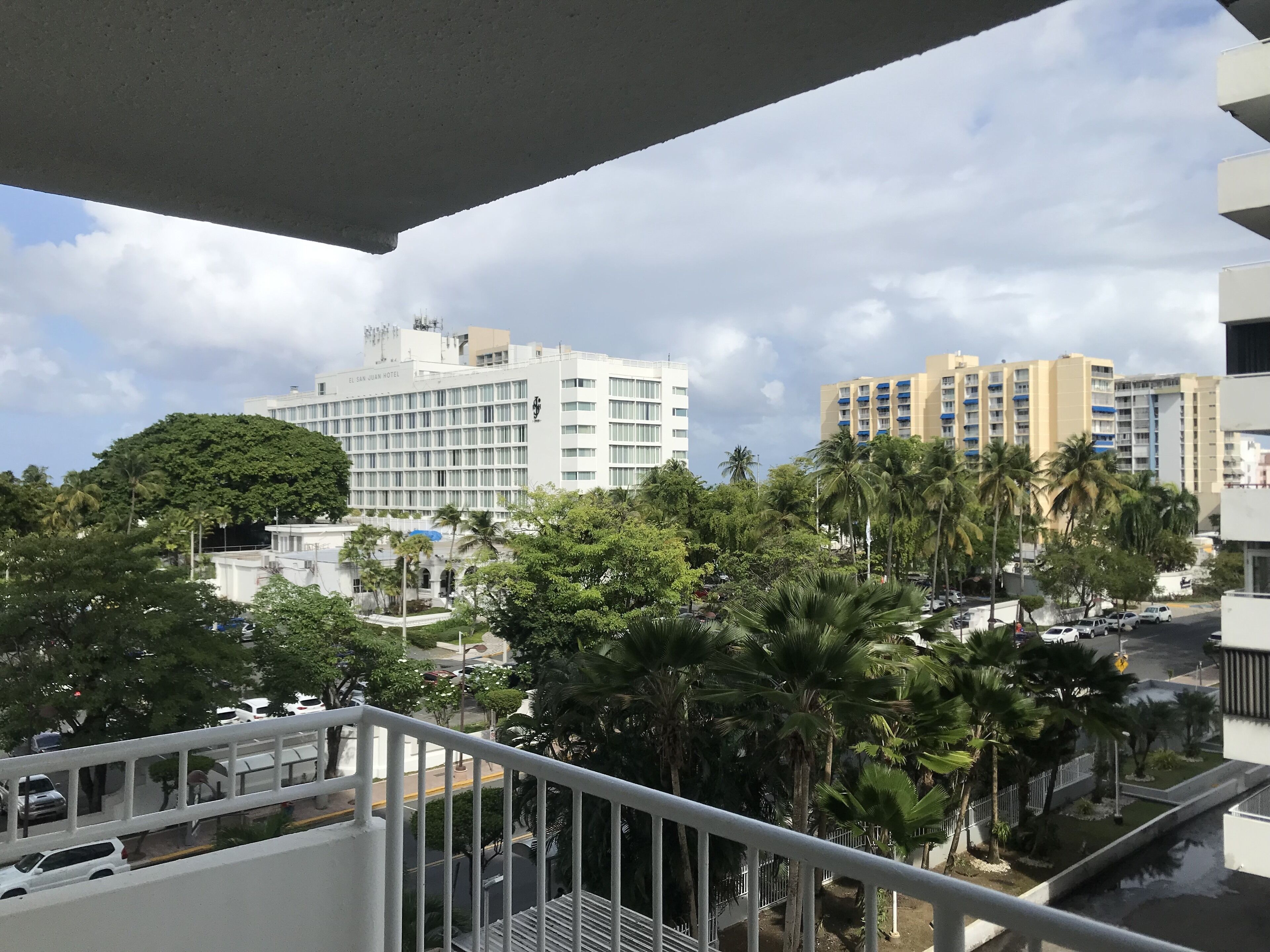 Un hôtel à étages, doté d’un balcon et offrant une vue sur les bâtiments environnants et les palmiers.