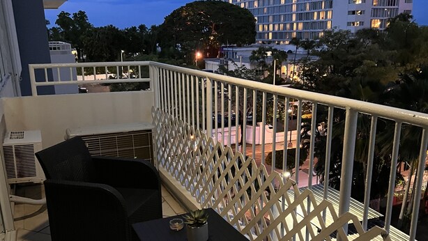 Un balcón con mobiliario exterior, barandilla de celosía blanca y vista de un edificio de hotel al fondo.