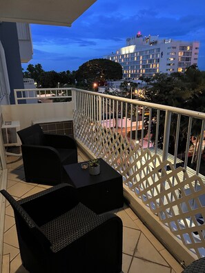 En balkon med udendørs møbler, et hvidt gittersgelænder og udsigt til en hotelbygning i baggrunden.