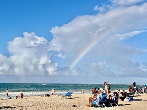 Ein Strand mit Menschen, die sich sonnen und schwimmen, einem klaren Himmel und einem deutlich sichtbaren Regenbogen.