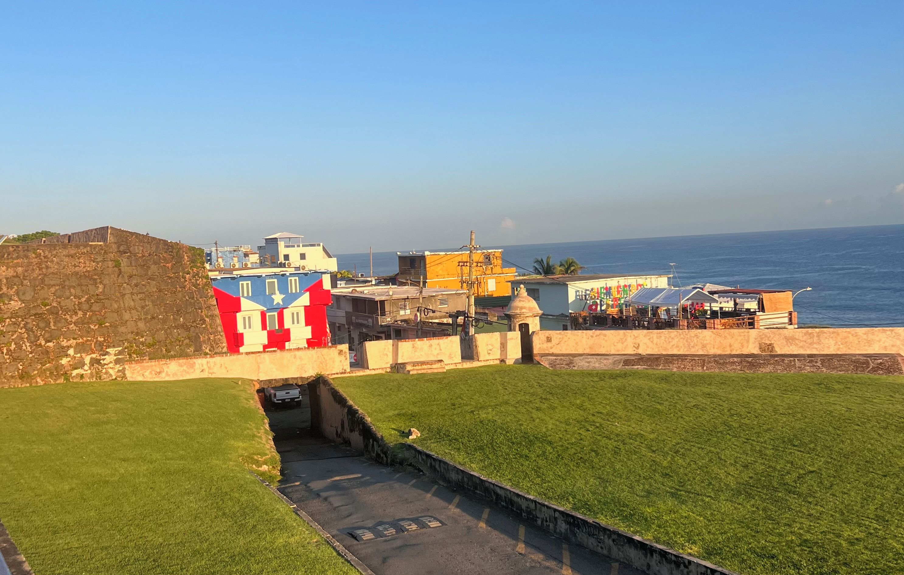 A coastal area with a stone wall, a grassy field, and buildings with colorful facades.