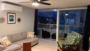 Un salón con un sofá gris, una mesa redonda naranja y un sillón estampado. Se ve un ventilador de techo, y la habitación tiene un balcón con vistas a edificios y árboles.