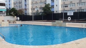 Una piscina con solárium de azulejos, rodeada por una valla y un edificio al fondo.