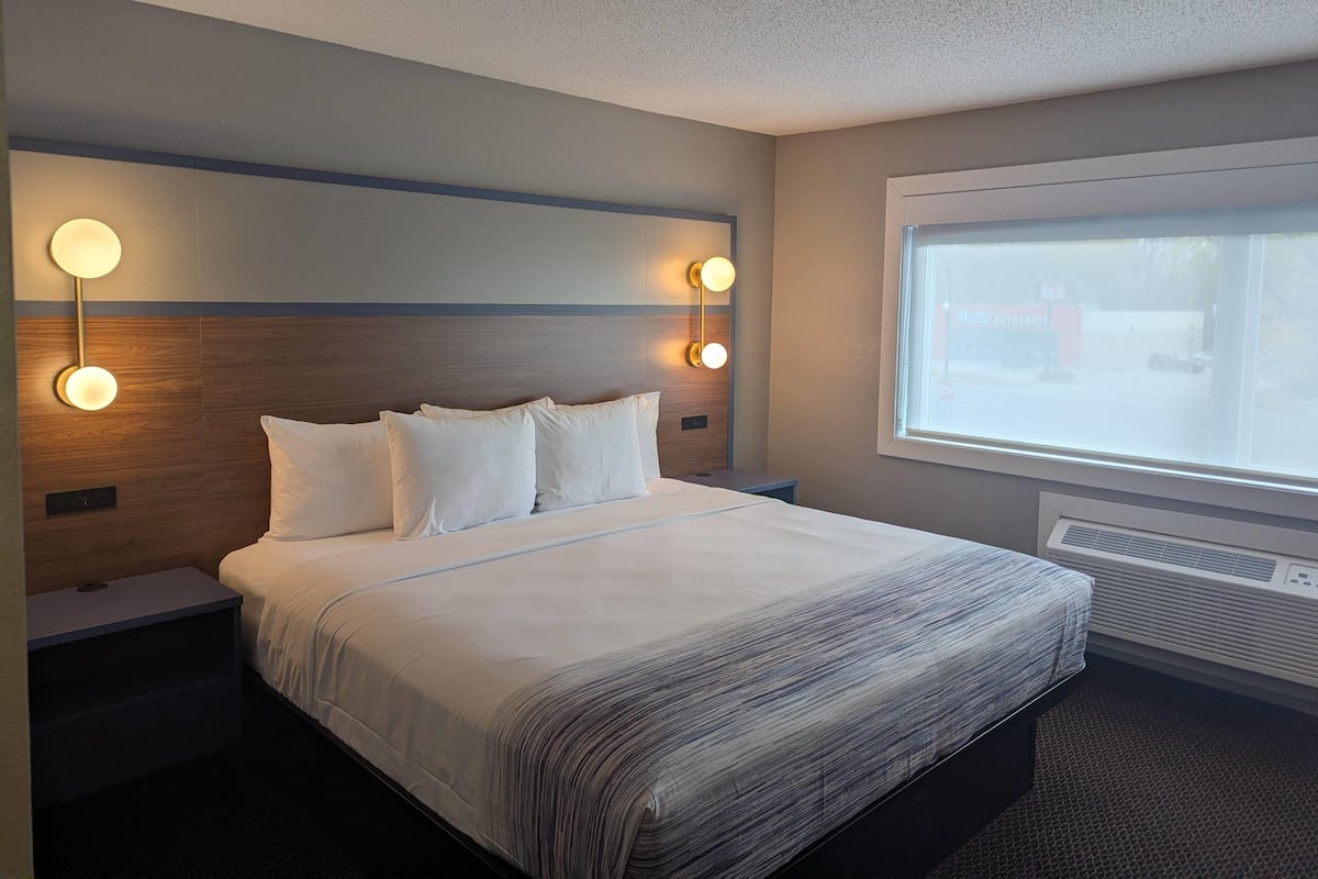 A hotel room with a large bed, two bedside lamps, a window with a view of the outside, and a wall-mounted air conditioning unit.