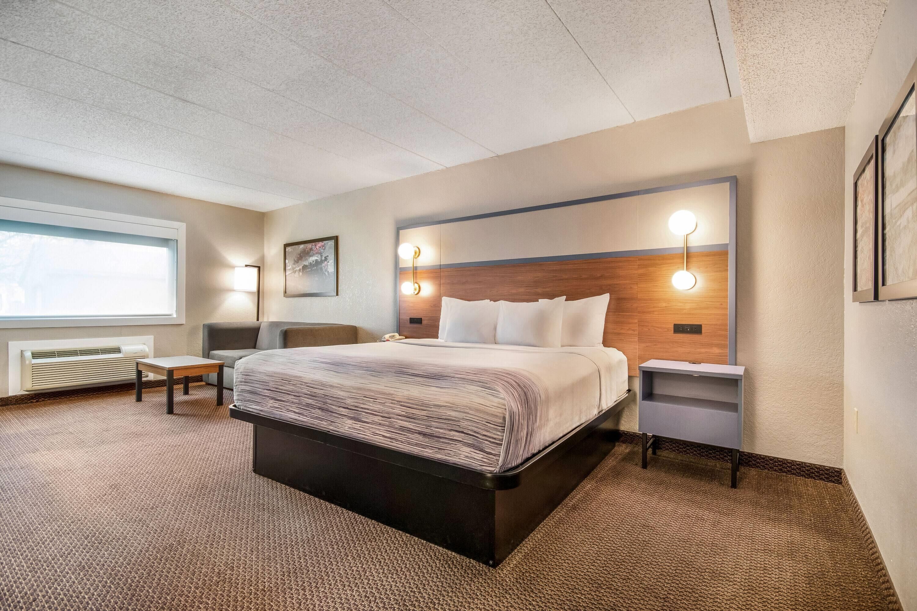 Une chambre d’hôtel avec un grand lit, des tables de chevet, une petite table, une fenêtre avec des stores et un luminaire fixé au mur.