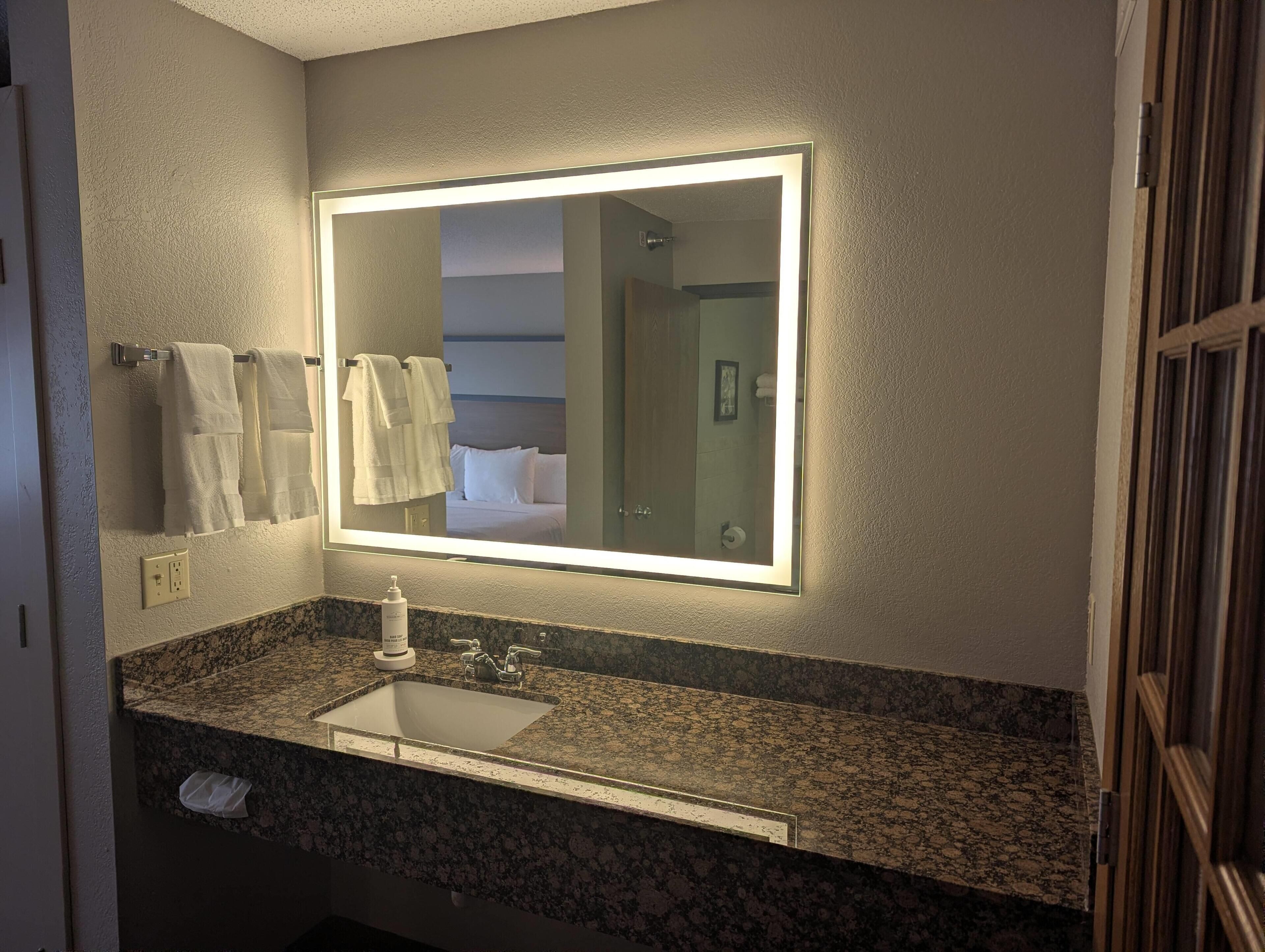 A bathroom with a granite countertop, a sink, a mirror with built-in lighting, and a towel rack with white towels.