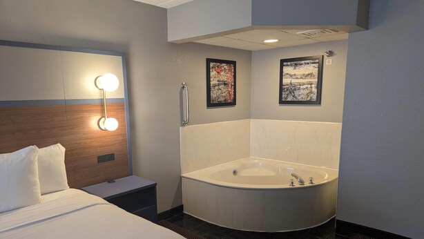 Habitación de hotel con una cama grande, una mesita de noche, una lámpara de pared, una bañera y dos cuadros enmarcados.