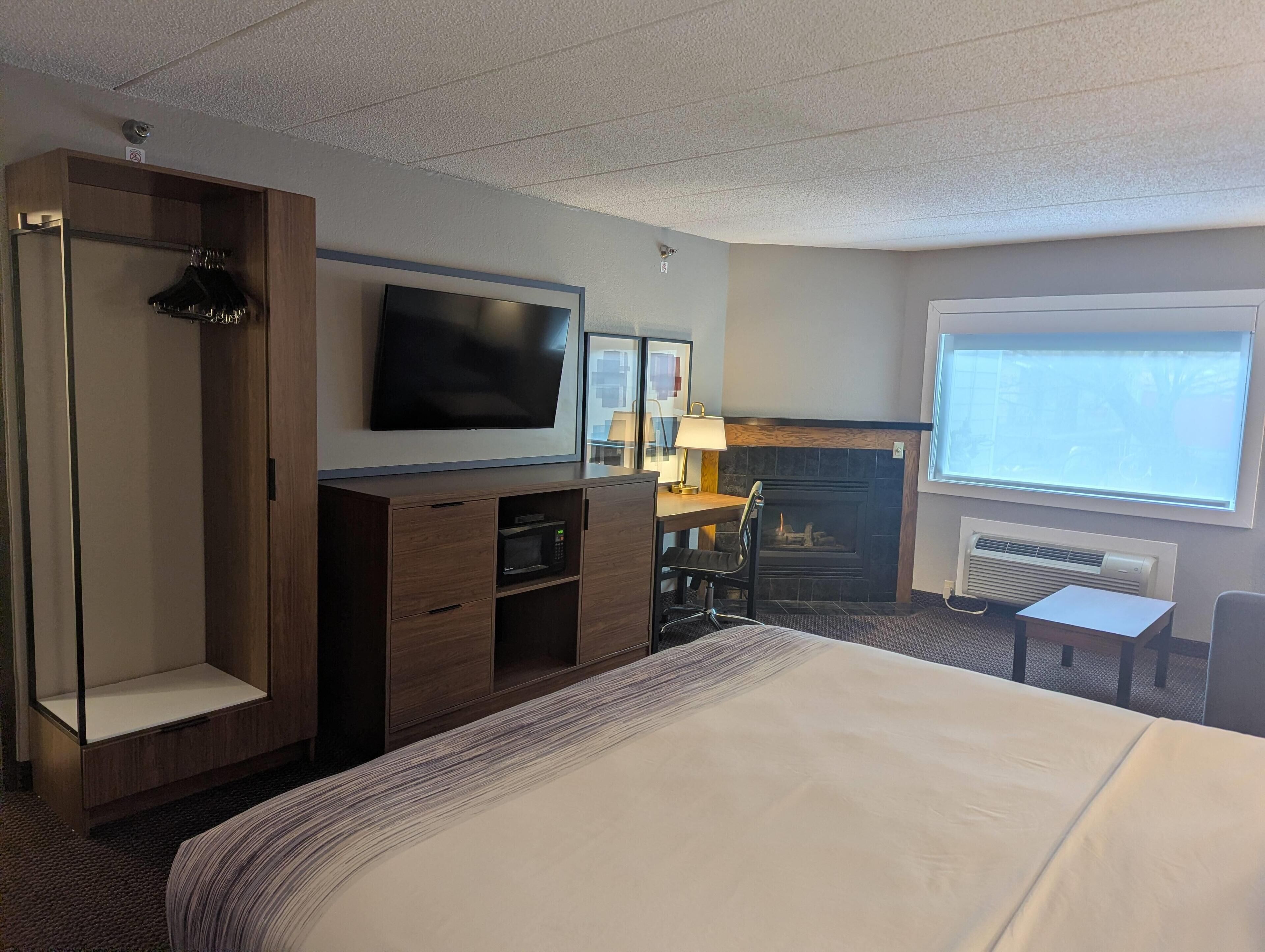 A hotel room with a bed, a TV, a desk with a lamp, a fireplace, and a window.