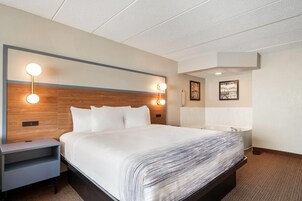 Una habitación de hotel con una cama grande, lámparas de noche, una mesita de noche, un baño con bañera y un cuadro en la pared.