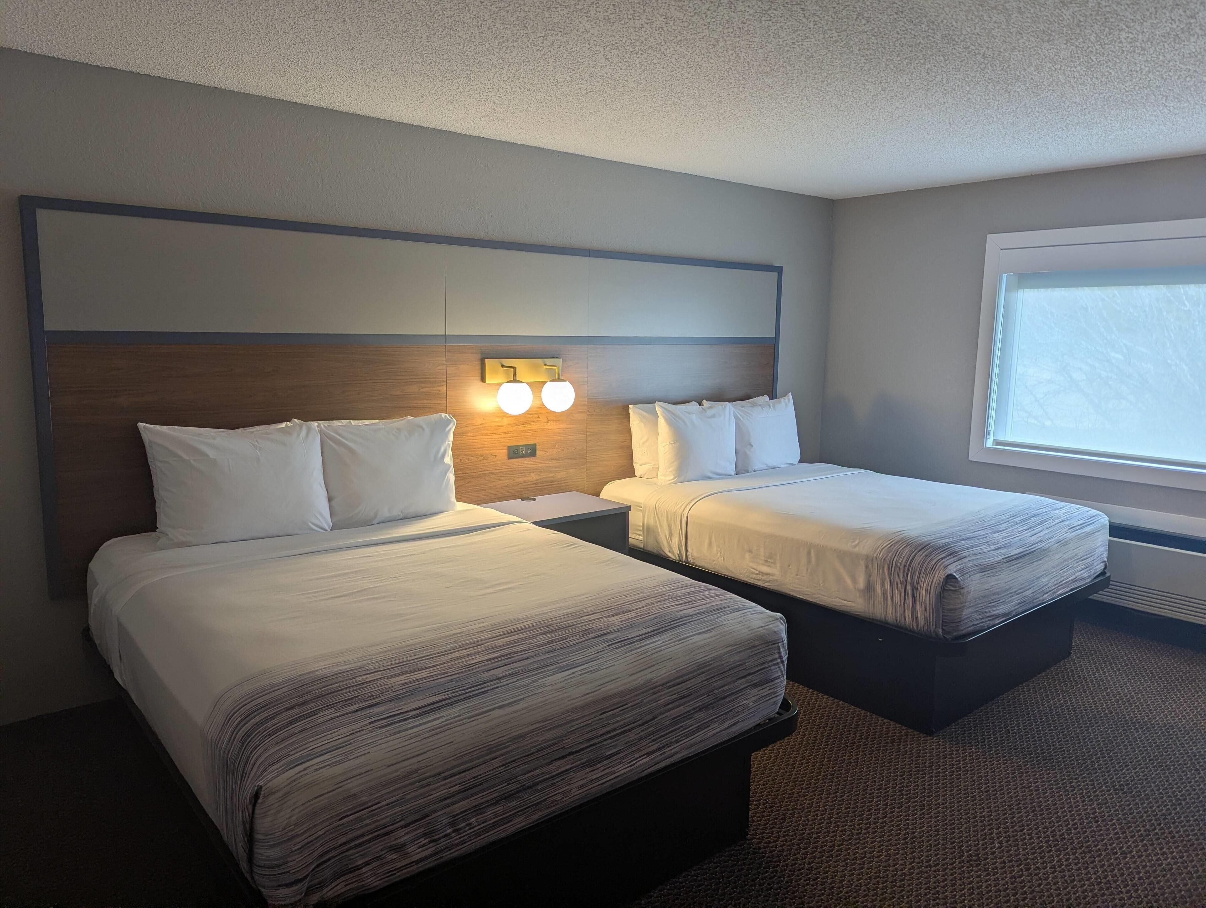 A hotel room with two beds, a wooden headboard, wall-mounted lights, and a large window.