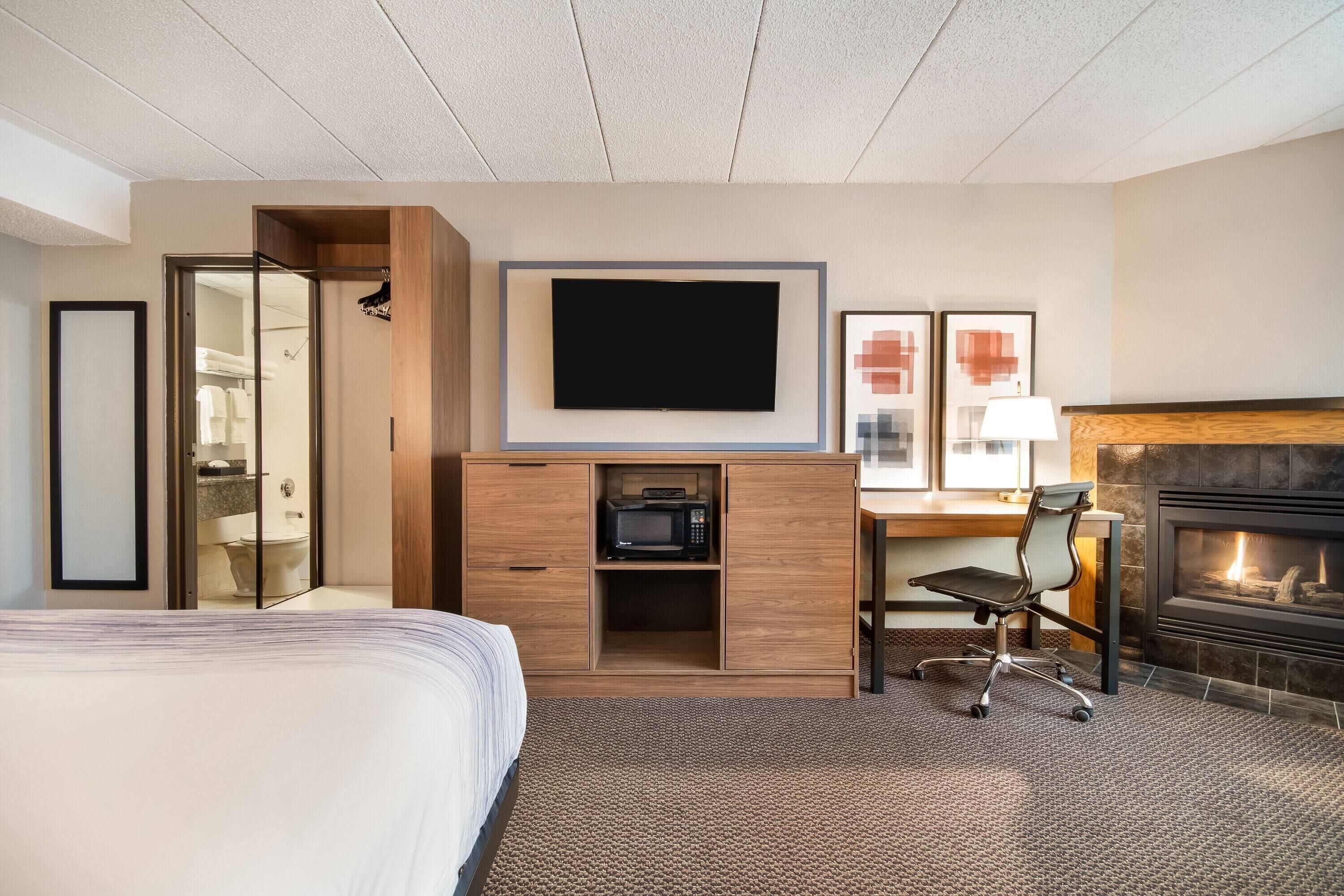 A hotel room with a bed, a TV mounted on the wall, a fireplace, a desk with a lamp, and a microwave in a wooden cabinet.