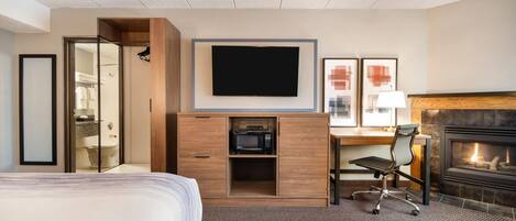 A hotel room with a bed, a TV mounted on the wall, a fireplace, a desk with a lamp, and a microwave in a wooden cabinet.