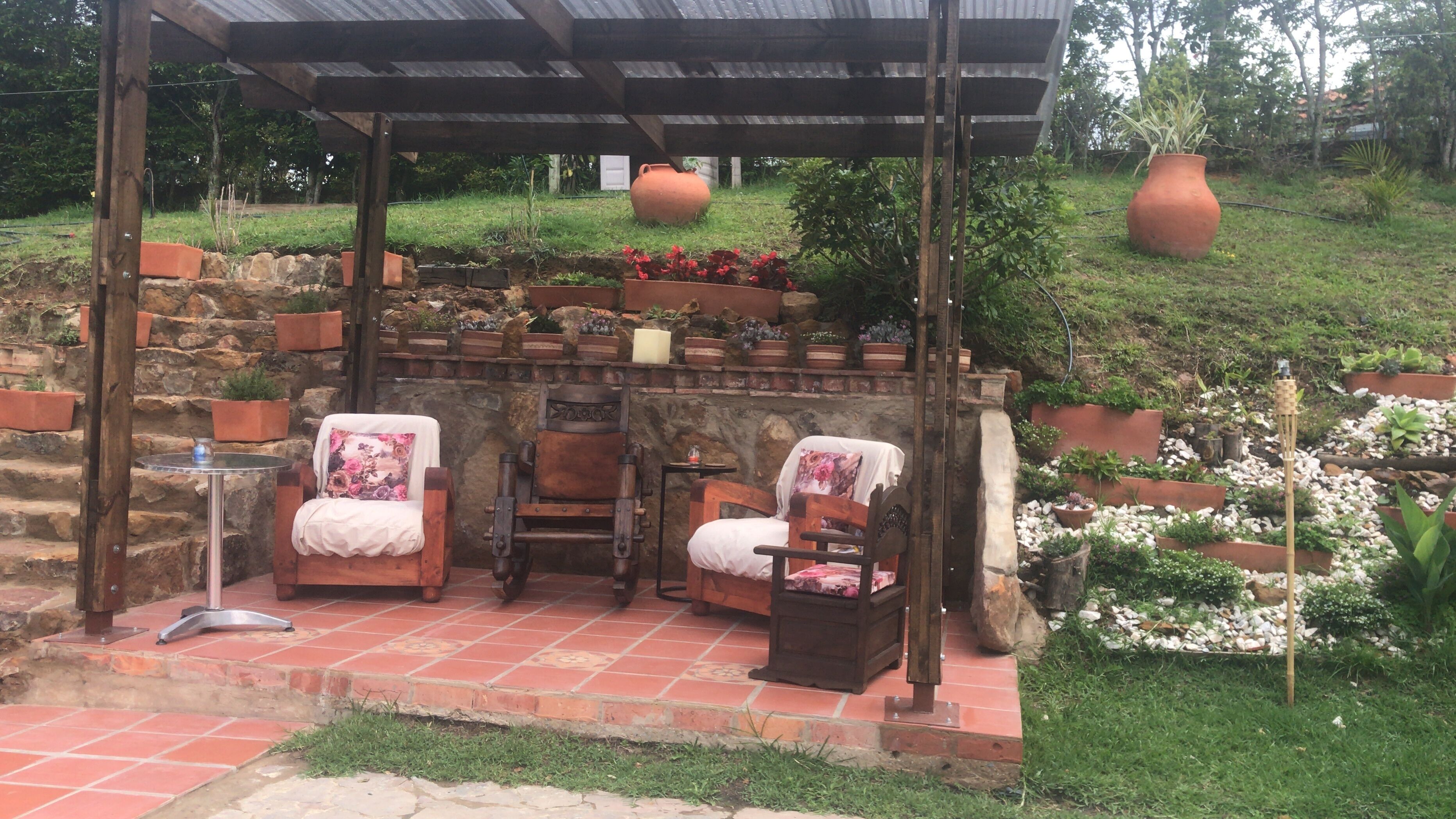A covered outdoor seating area with two wooden chairs, a small table, and a stone wall with potted plants.