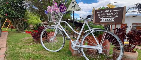 Una bicicleta blanca con una cesta llena de flores estacionada en un jardín, con un cartel que dice "Hoy es un buen día para ser feliz".