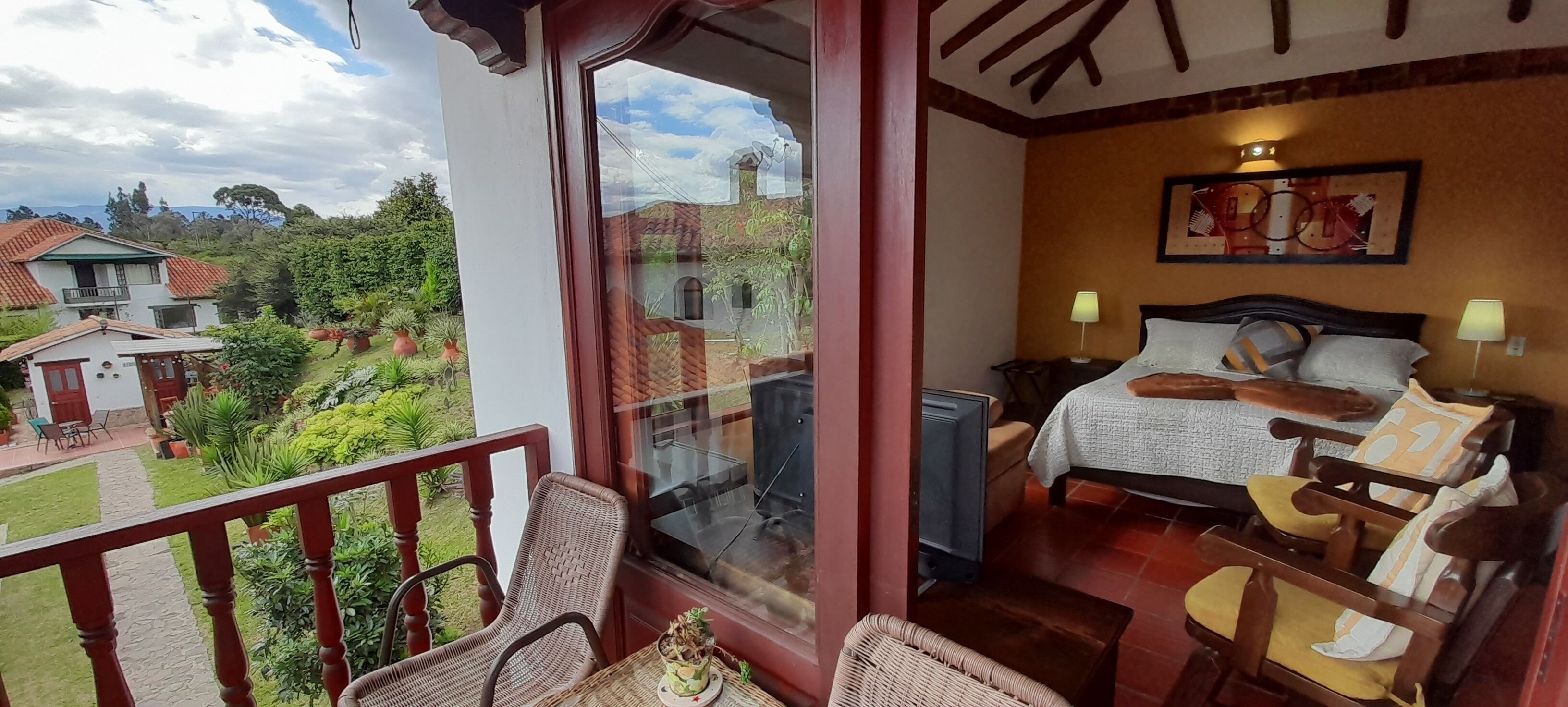 A room with a bed, two bedside tables, a window with a view of a garden and buildings, and a balcony with wicker furniture.
