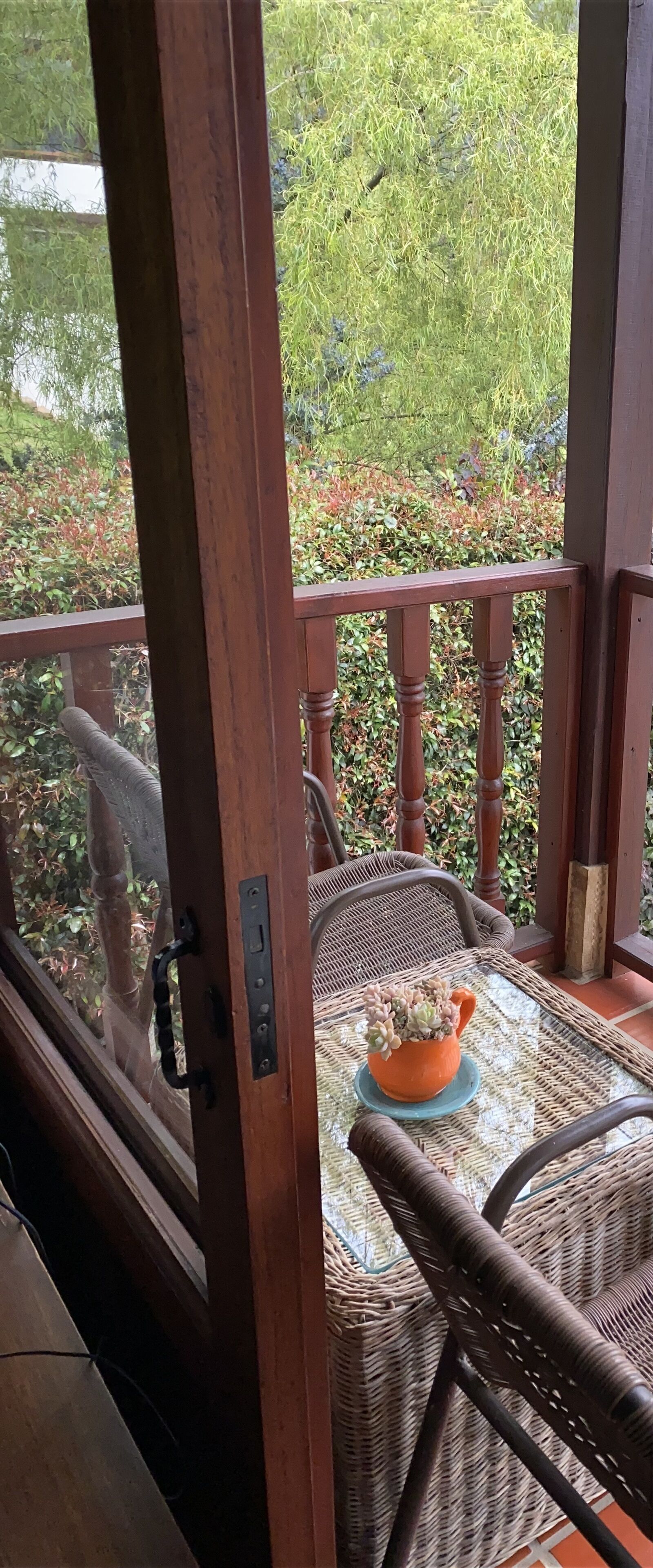 Un balcón de madera con una mesa y sillas de mimbre, una planta en maceta y vistas a un jardín.