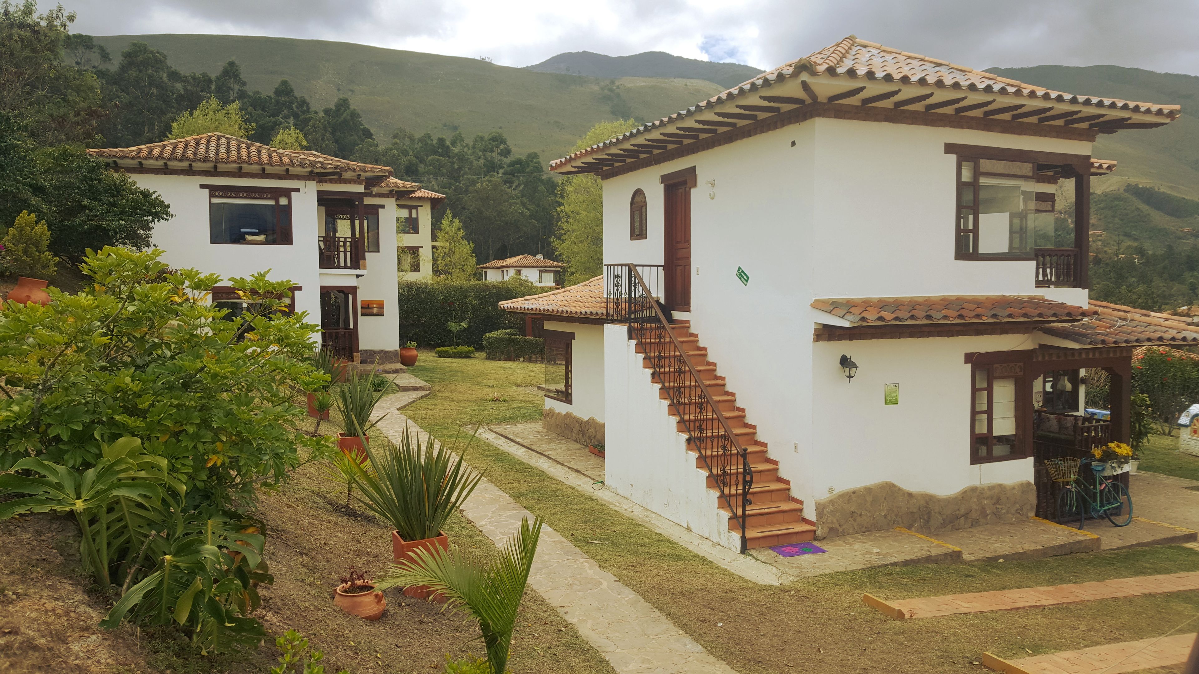 Un edificio de dos pisos con una escalera que conduce al nivel superior, rodeado de abundante vegetación y colinas al fondo.