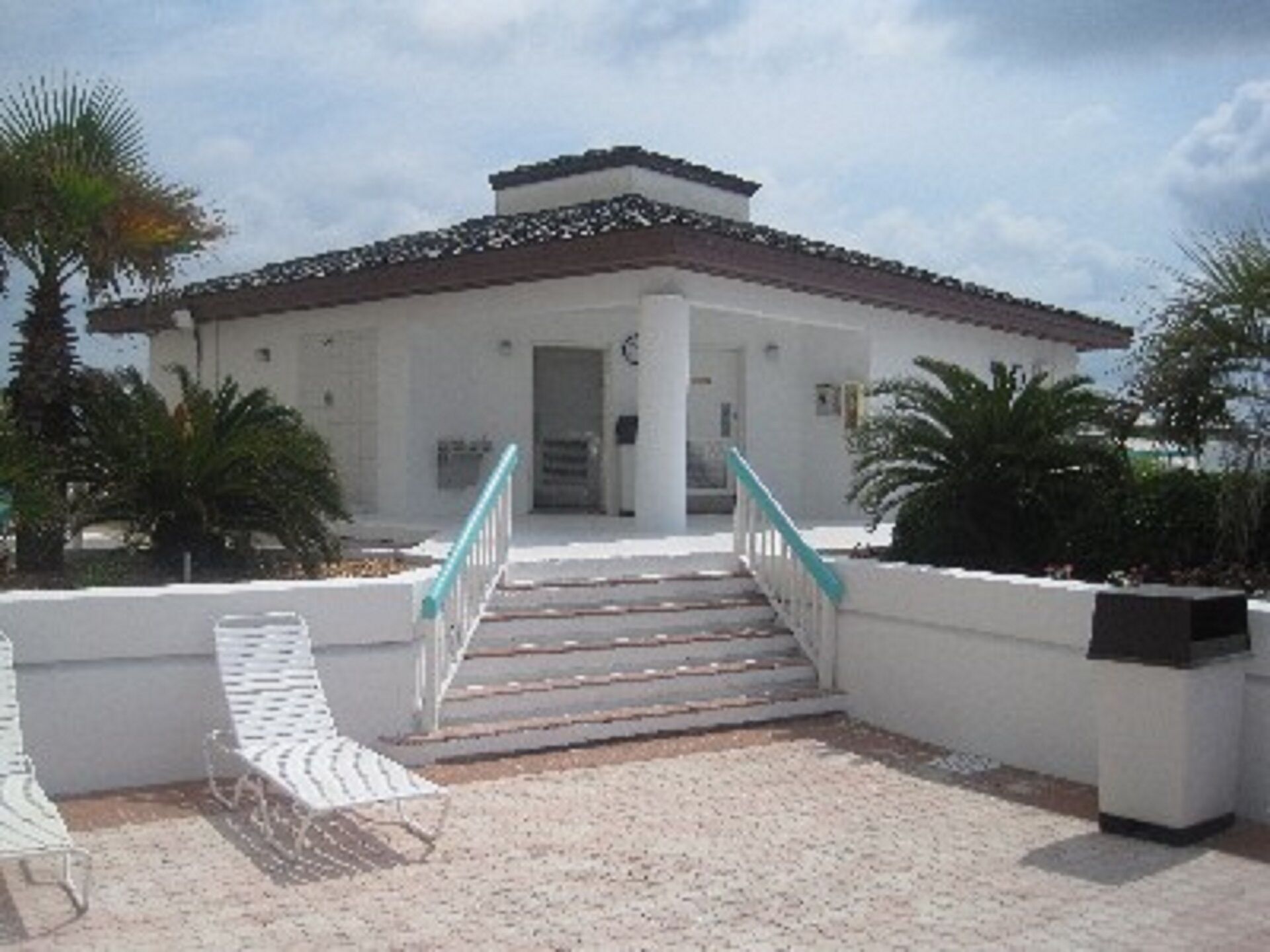 A building with a white facade, a tiled roof, and a staircase leading to the entrance. There are palm trees and a bench in the foreground.