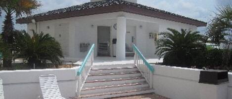 A building with a white facade, a tiled roof, and a staircase leading to the entrance. There are palm trees and a bench in the foreground.