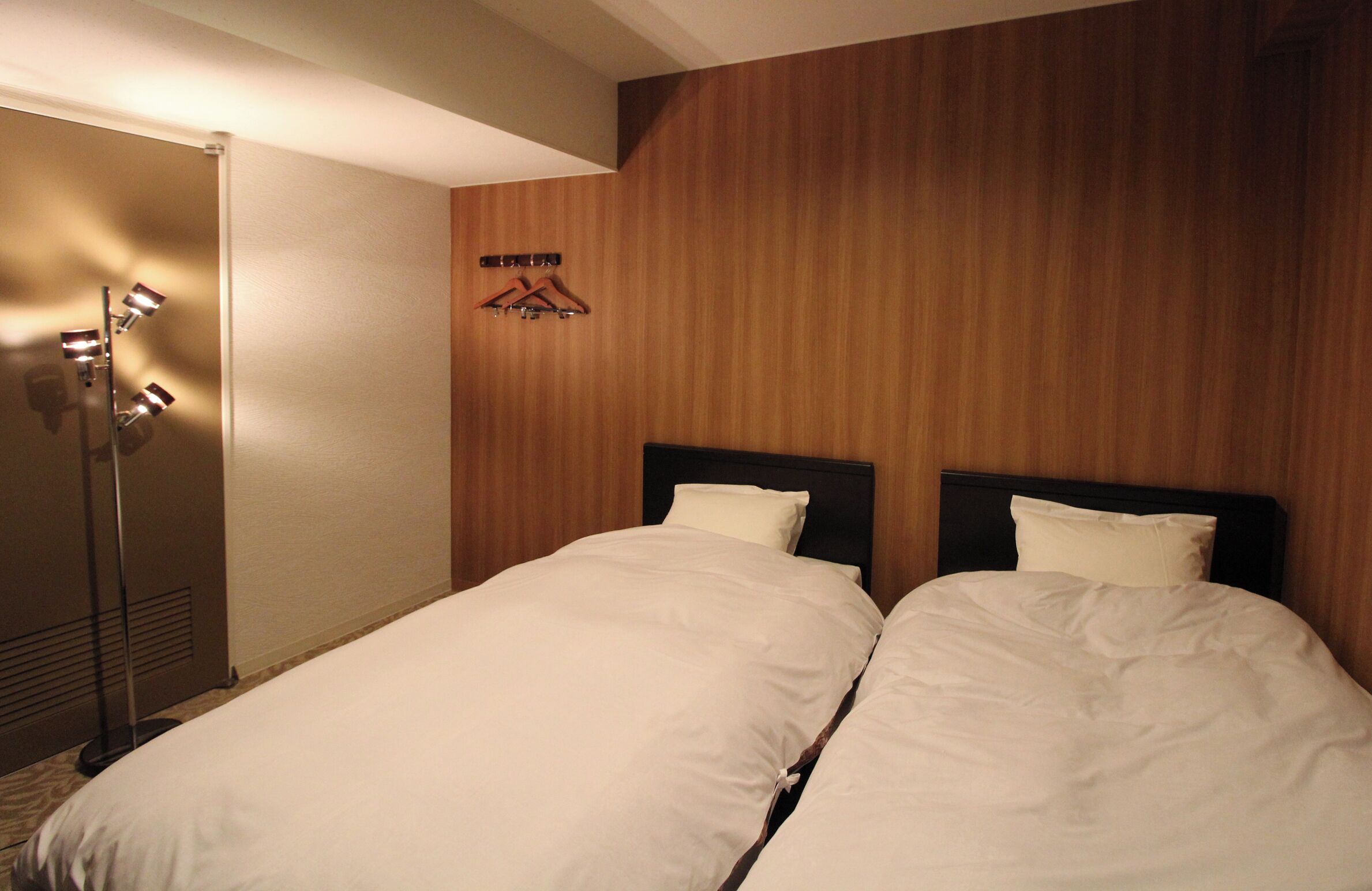 A hotel room with two beds, a wooden wall, a coat rack, and a floor lamp.