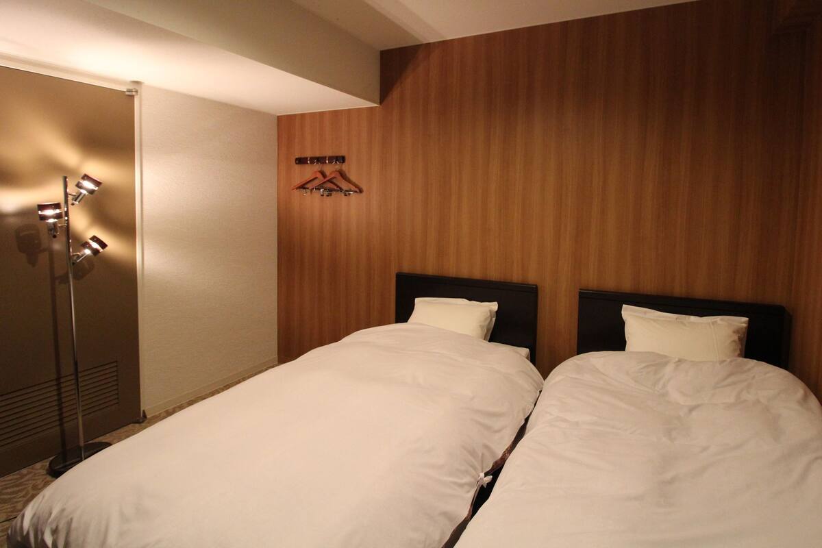A hotel room with two beds, a wooden wall, a coat rack, and a floor lamp.