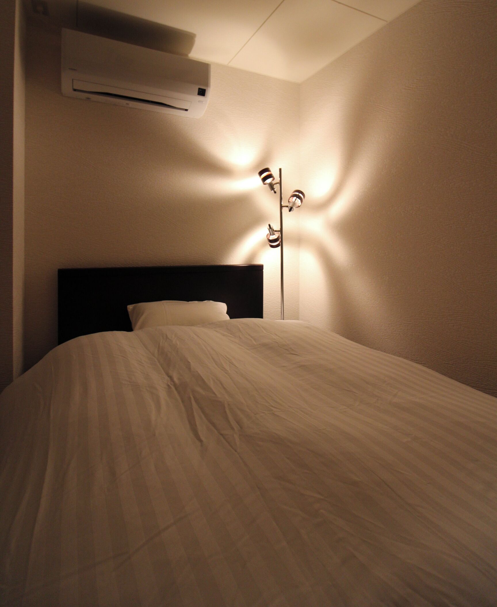 A bed with a headboard, an air conditioning unit above it, and a floor lamp casting shadows on the wall.