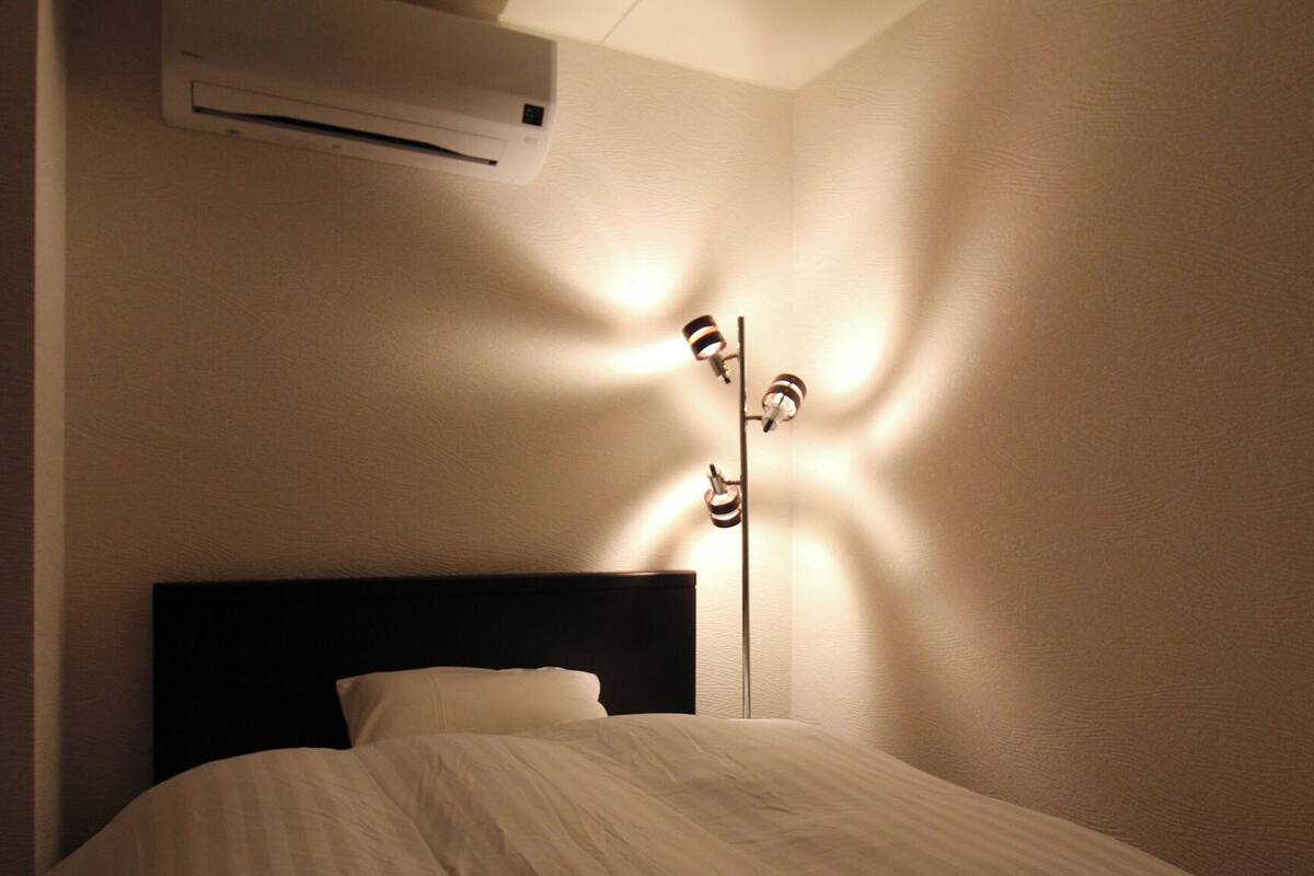A bed with a headboard, an air conditioning unit above it, and a floor lamp casting shadows on the wall.