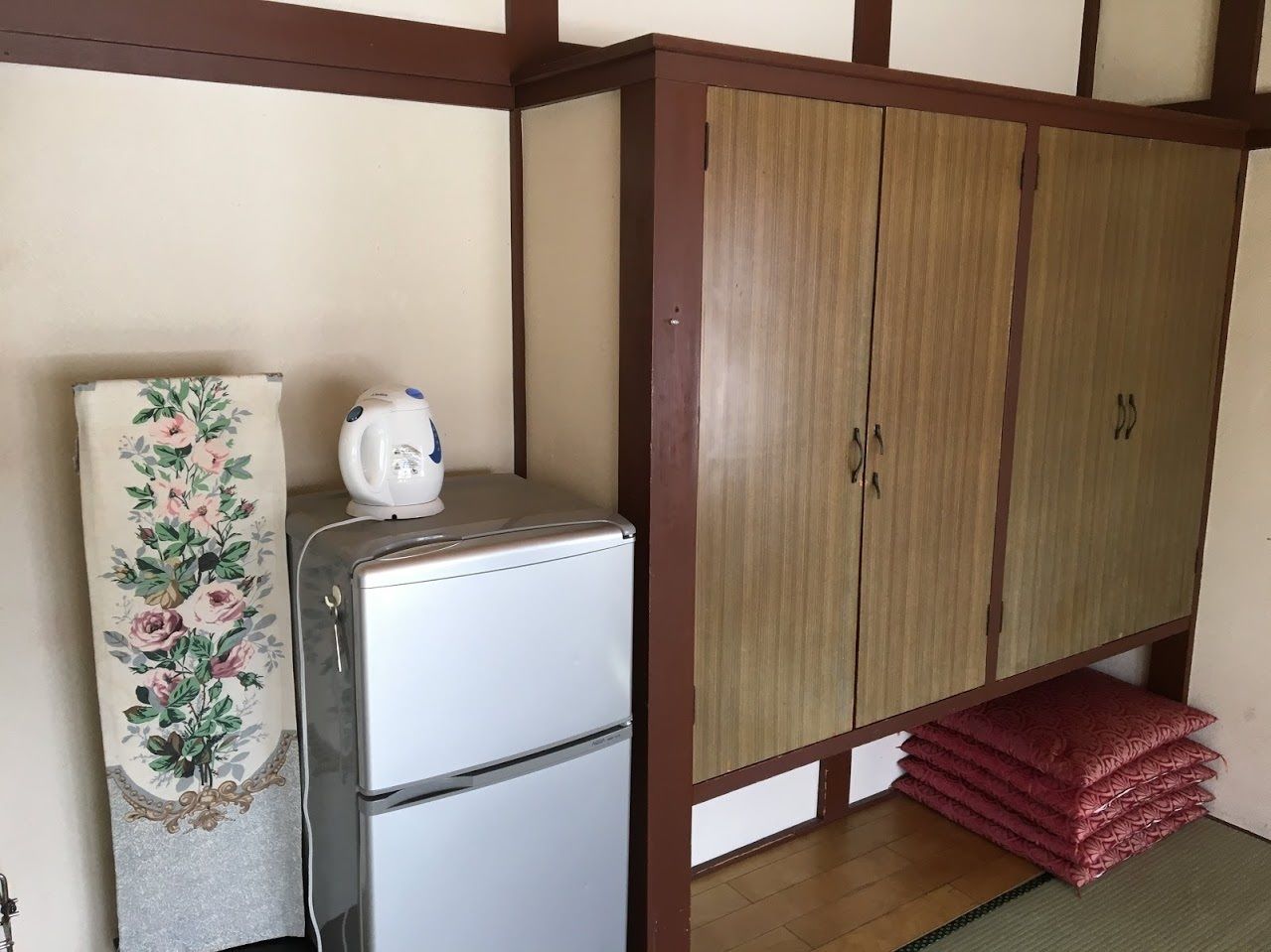 A compact room with a kitchenette area, including a refrigerator and a microwave, and a wardrobe with a wooden finish.