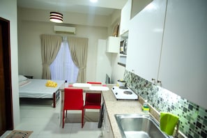 A compact hotel room with a kitchenette, a bed with curtains, a red chair, and a small dining table.