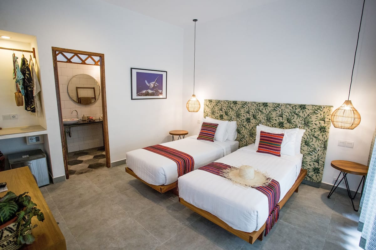 Habitación de hotel con dos camas, una kitchenette pequeña, un baño con espejo redondo y vista al mar.