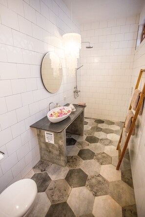 Un baño con paredes de azulejos blancos, piso con diseño hexagonal, espejo redondo, lavamanos con cuenco y escalera de madera.