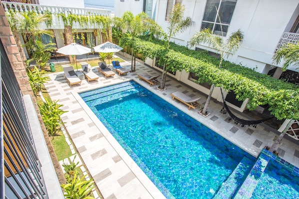 Une piscine sur le toit, agrémentée de chaises longues et de parasols, entourée de verdure et bordée d’un bâtiment blanc.