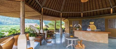A spacious, open-air lounge with wicker furniture, a reception desk, and a view of the surrounding landscape.