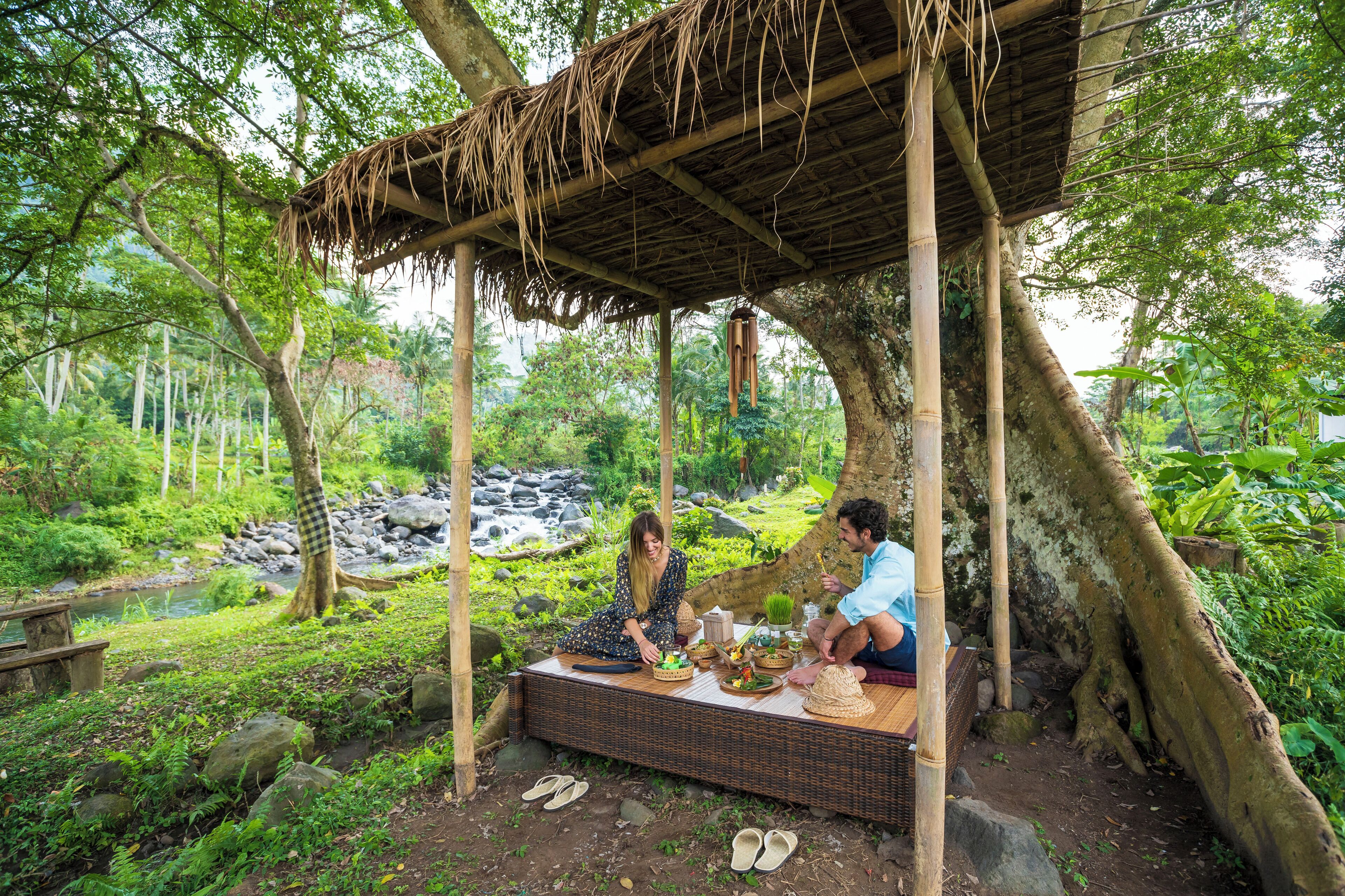 A rustic outdoor dining area with a thatched roof, surrounded by lush greenery and a small stream.