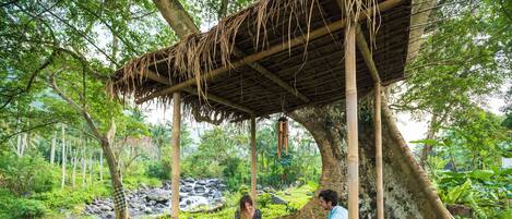 A rustic outdoor dining area with a thatched roof, surrounded by lush greenery and a small stream.