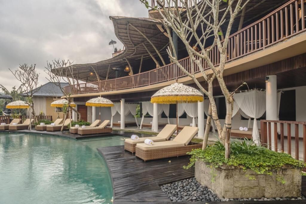 Zona de piscina con sillones de descanso y sombrillas, terraza de madera y un edificio con balcón.