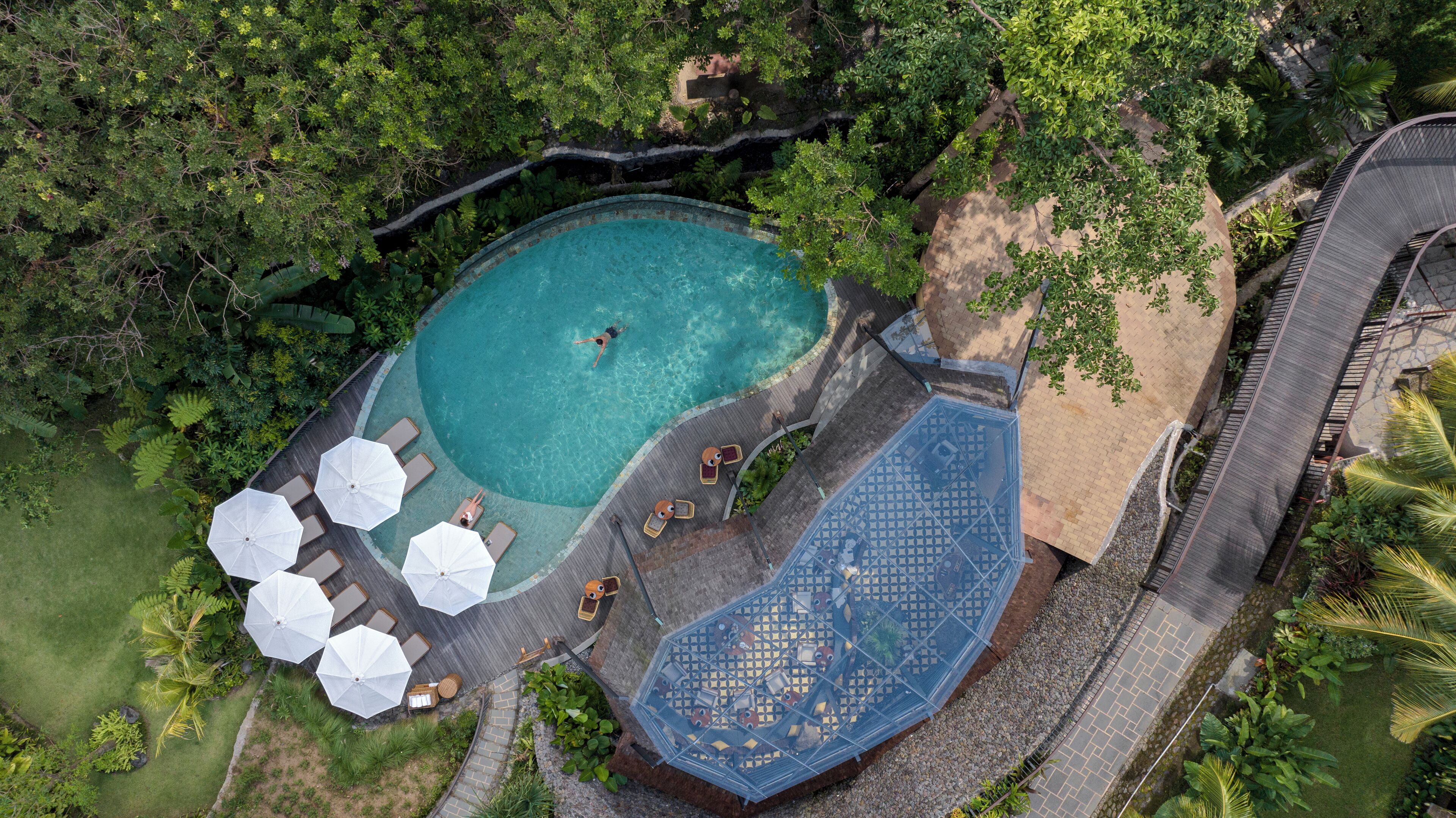 An aerial view of a resort with a swimming pool surrounded by lounge chairs and umbrellas, a covered seating area, and lush greenery.