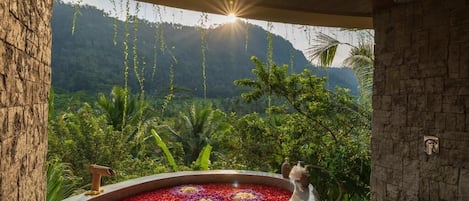Un área de spa con una bañera de hidromasaje rodeada de flores rojas y púrpuras, con un fondo de exuberante vegetación y montañas.