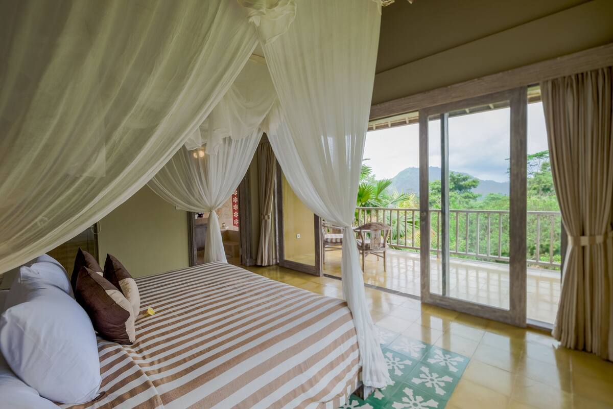 Una habitación con cama, cortinas blancas, un balcón con vistas a la vegetación y montañas.
