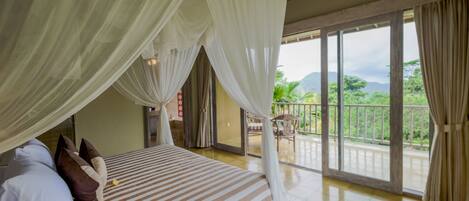 A room with a bed, white curtains, a balcony with a view of greenery and mountains.