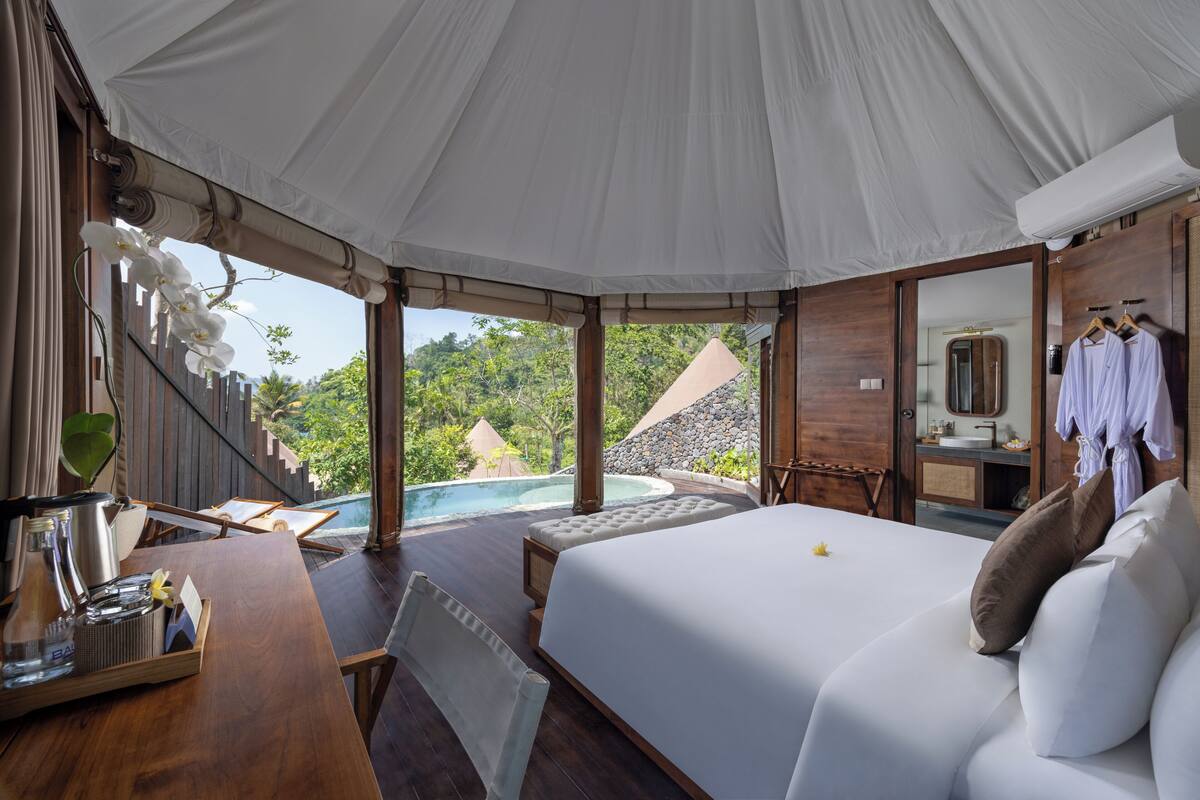 Un dormitorio con una cama grande, un escritorio de madera con bandeja, vistas a una piscina y un baño con espejo y perchero para bata.