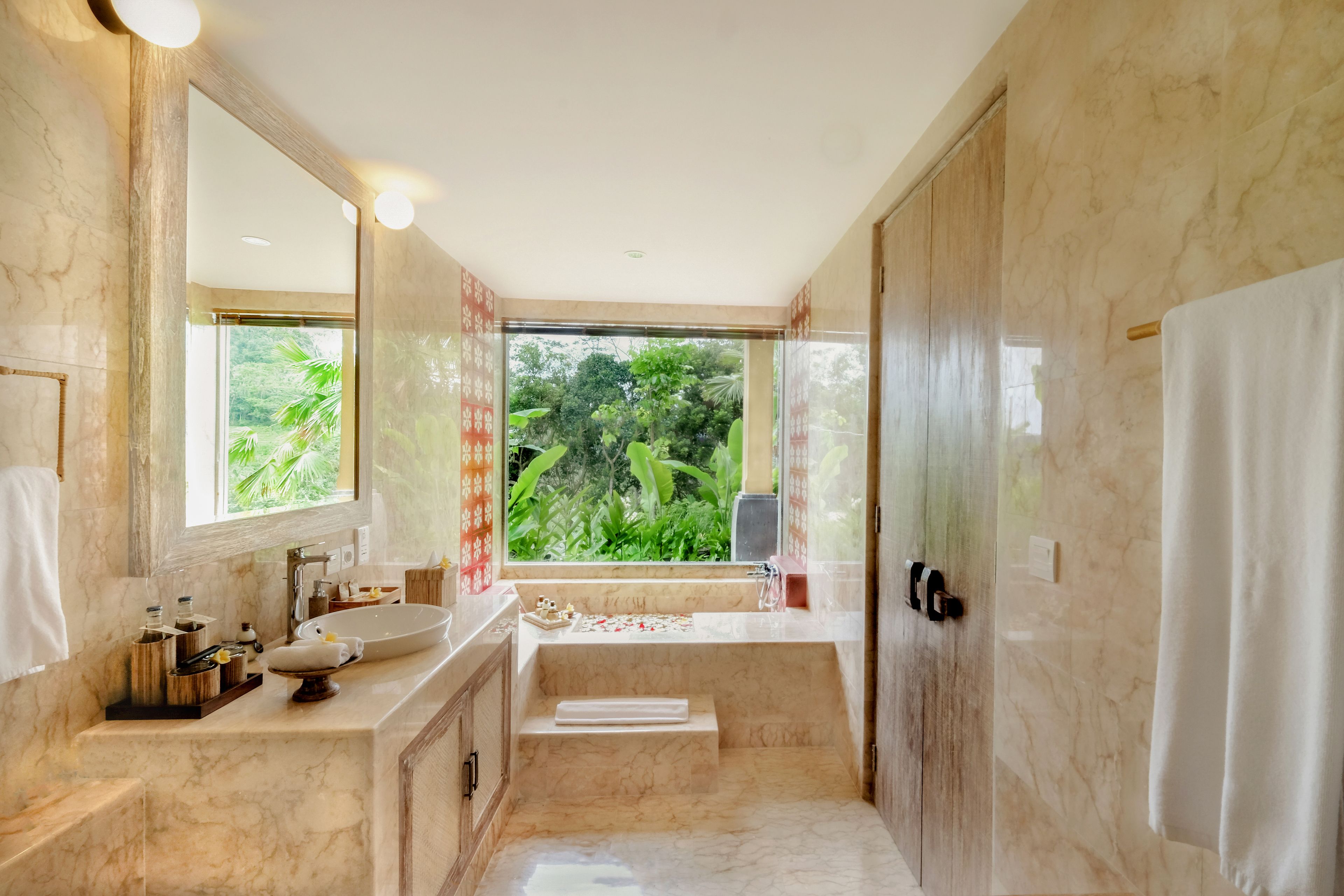 A modern bathroom with a large sink, mirror, and bathtub, featuring marble walls and a view of greenery.