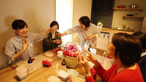 A group of people sitting around a wooden table in a room with a kitchen in the background. - COCORETO THE GUEST HOUSE - Hostel (Matsue)