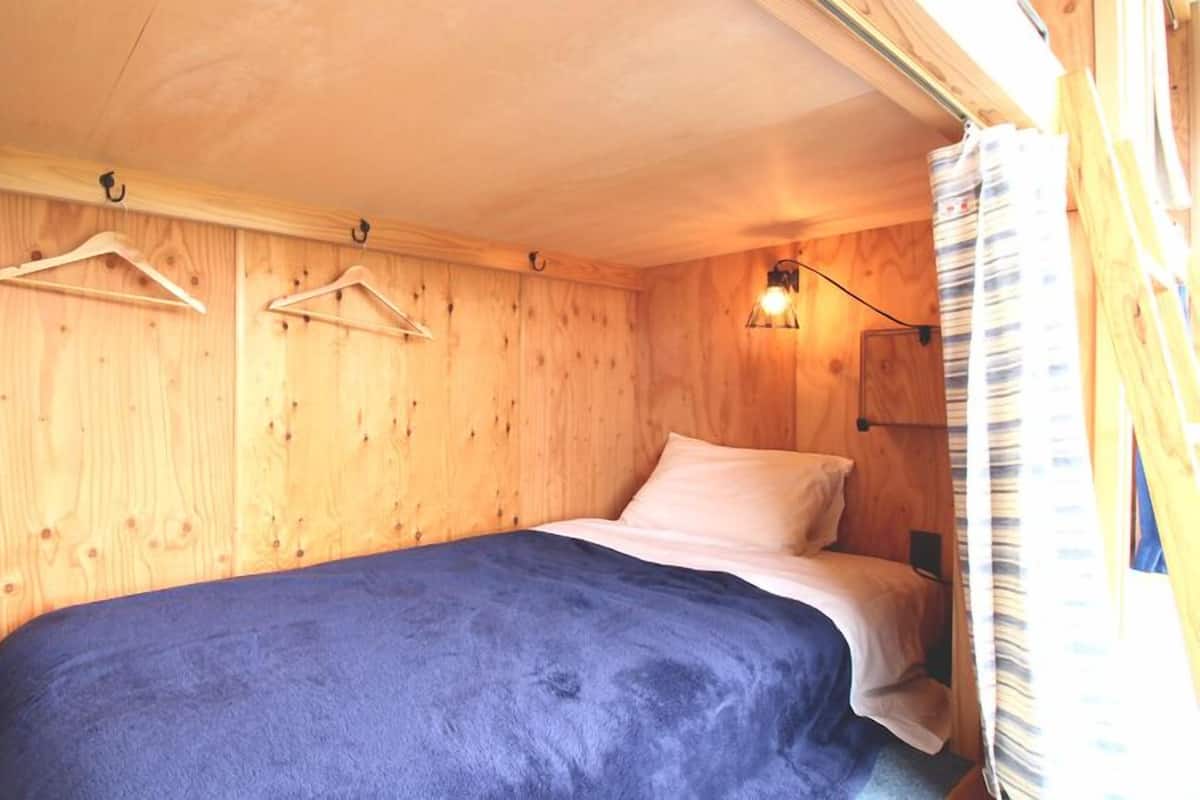 A small, wooden room with a bed, a hanging light fixture, and a window with curtains.