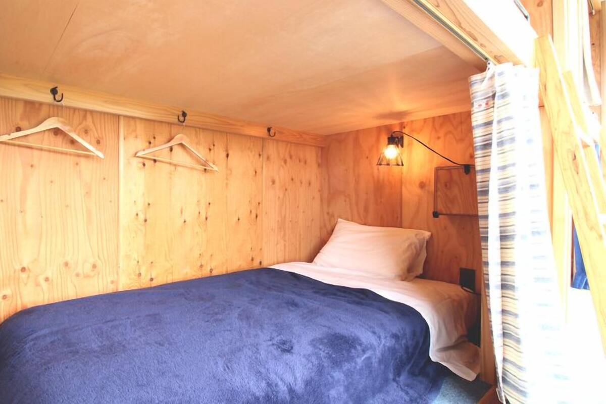 A small, wooden room with a bed, a hanging light fixture, and a window with curtains.