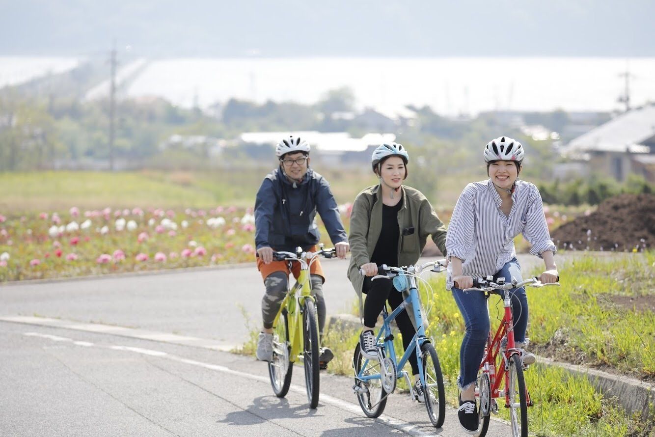 Three people riding bicycles on a paved path with flowers on the side.